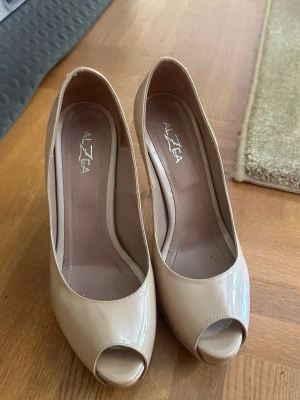 Beige peep toe pumps från Alzea - Snygga beige pumps från Alzea med hög klack och peep toe-design. Skorna är i glansigt lackmaterial som ger en elegant look. 2000 talets mest trendigaste design! 