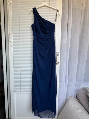 Marinblå oneshoulder långklänning - Elegant marinblå långklänning med oneshoulder-design. Klänningen har ett draperat liv och faller snyggt längs kroppen. Perfekt för speciella tillfällen där du vill sticka ut med en stilren look. Ny med prislapp, aldrig använd.