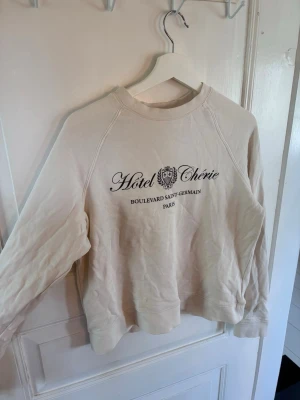 Beige sweatshirt från Holly & Whyte - Beige sweatshirt från HOLLY & WHYTE med trycket 'Hotel Chérie Boulevard Saint-Germain Paris' i svart framtill. Tröjan har rund halsringning, långa ärmar och ribbade muddar. Tillverkad i mjukt bomullsmaterial för en avslappnad look.