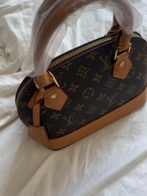 Väska - Snygg handväska i klassiskt monogrammönster. Väskan är brun med ljusbruna detaljer och guldfärgade metalldelar. Har rundad form samt dragkedja upptill.