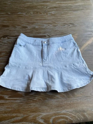 Ljusblå volangkjol med jeanslook - Söt ljusblå kortkjol med volangkant och klassisk jeanslook. Kjolen har fickor fram, knapp och dragkedja i midjan. Perfekt för dig som gillar en avslappnad men trendig stil.