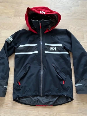 Blå röd Helly Hansen seglar/regnjacka - Svart regnjacka från Helly Hansen med röd huva och grå fleecefoder. Jackan har vita reflexdetaljer, två snedställda fickor med dragkedja och justerbara muddar. Perfekt för blöta dagar och outdoor-äventyr.