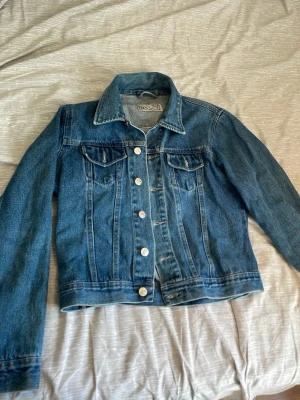 Klassisk blå jeansjacka - Snygg blå jeansjacka till tjejer i klassisk modell med raka linjer och silverfärgade knappar framtill. Jackan har två bröstfickor med lock och knapp samt långa ärmar. Perfekt att slänga över en hoodie eller t-shirt för en avslappnad look aldrig använd! Hör av er vid funderingar!