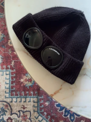 Svart mössa med goggles från C.P. Company - Säljer en svart ribbstickad mössa från C.P. Company med ikoniska goggles på uppviket. Mössan är tillverkad i mjukt material och har en modern, streetinspirerad look som sticker ut. Perfekt accessoar för dig som gillar unik stil.