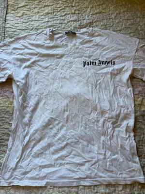 Vit Palm Angels t-shirt - Säljer en vit t-shirt från Palm Angels med svart logga tryckt på bröstet och stort tryck på ryggen. Klassisk passform och rund hals. Perfekt för dig som gillar streetwear och vill ha en enkel men snygg look.