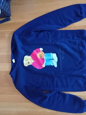 Mörkblå sweatshirt med Polo Bear - Säljer en mörkblå sweatshirt från Ralph Lauren med den ikoniska Polo Bear-trycket på framsidan. Björnen har röd tröja, ljusblå jeans och håller en basketboll. Tröjan har rund hals och långa ärmar, perfekt för en avslappnad streetwear-look.