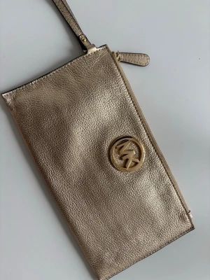 Guldig kuvertväska Michael Kors - Snygg kuvertväska i metallic guld från Michael Kors med MK-logga framtill. Väskan har dragkedja upptill och en smidig handledsrem. Tillverkad i glansigt läder som ger en lyxig känsla och passar perfekt när du vill sticka ut. 