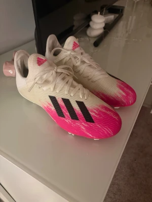 Adidas vita och rosa fotbollsskor - Snygga fotbollsskor från Adidas i vitt med rosa detaljer och svarta ränder på sidan. Skorna har snörning och perfekt för snabba rörelser på planen. Tillverkade i syntetmaterial för bra grepp och komfort. Endast använda 2 gånger.