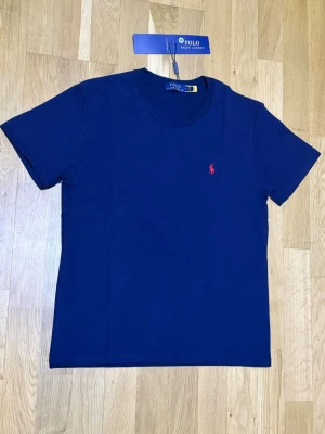 Marinblå t-shirt från Polo Ralph Lauren - Snygg marinblå t-shirt från Polo Ralph Lauren med klassisk passform och korta ärmar. T-shirten har en liten röd broderad logga på bröstet och är tillverkad i mjuk bomull. Perfekt för en stilren och avslappnad look.
