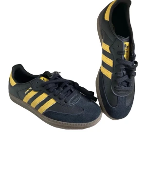Adidas Samba svart/gul sneakers - Helt nya Adidas Samba sneakers i svart med gula detaljer och klassiska ränder på sidan. Skorna har en ovandel inlocka och läder, samt gummisula. Perfekt för den som gillar en retrostil och streetwear då de inte längre tillverkas!