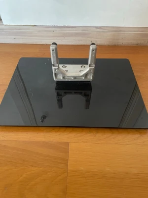 Svart glasbas till TV-stativ - Säljer en rektangulär svart glasbas med metallfäste, perfekt för att montera en TV eller skärm. Basen är i glas och fästet är i silverfärgad metall. Passar till olika TV-modeller och ger stabilitet på plana ytor.