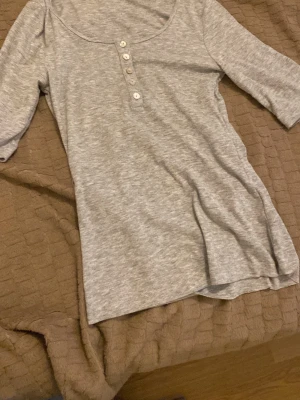 Grå ribbad topp med knappar - Grå ribbad topp med trekvartsärm och rund halsringning. Toppen har knappar framtill och är tillverkad i ett mjukt, stretchigt material som sitter skönt på kroppen. Perfekt att styla med jeans eller kjol.