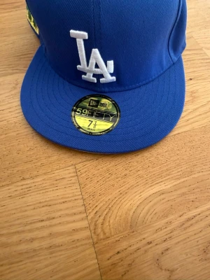 Blå LA keps med broderi - Snygg blå keps från New Era med LA-logga framtill och broderade palmer och text på sidan. Klassisk flat peak och detaljer i vitt, grönt och gult. Perfekt för dig som vill sticka ut med en unik accessoar.