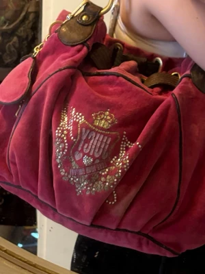 Juicy Couture röd velourväska  - Cool röd handväska i velour med bruna detaljer och guldfärgade metalldelar. Väskan har ett stort emblem framtill dekorerat med strass och tryck, samt dragskoöppning. Perfekt för dig som gillar Y2K-stil och vill sticka ut.