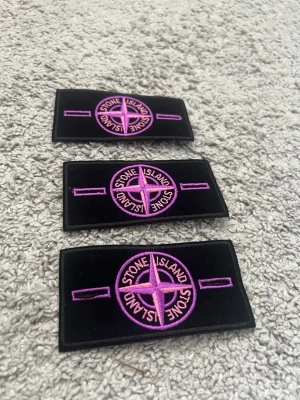 Stone Island patchar svart/lila 3 styckna - Tre stycken svarta tygpatchar från Stone Island med broderad kompasslogga i lila. Rektangulär form och tydliga detaljer, perfekta att sy eller fästa på jackor, väskor eller andra plagg för en unik look.