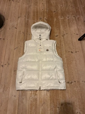 Vit dunväst med huva från Moncler - Snygg vit dunväst från Moncler med huva och dragkedja framtill. Västen har två fickor med dragkedja och Moncler-logga på bröstet. Perfekt för lager på lager och håller dig varm under kyliga dagar.