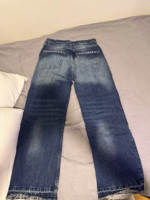 Blå jeans med pärldetaljer bak - Snygga blå jeans med raka ben och slitna kanter nertill. Baktill finns två fickor med pärldetaljer längs kanten som ger en cool touch. Jeansen har en klassisk femficksmodell och är tillverkade i denim.
