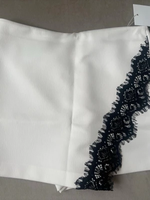 Viral byxkjol med spets från ZARA - Snygga vita shorts från ZARA med en dekorativ svart spetsdetalj längs ena sidan. De har en stilren design med dold dragkedja framtill och är tillverkade i ett slätt tyg som känns lyxigt. Perfekta för dig som vill sticka ut med en trendig look.