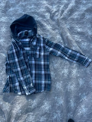 Rutig overshirt med huva från DC - Säljer en snygg rutig overshirt från DC i blått, vitt och svart. Skjortan har lång ärm, bröstficka och en mörkblå huva i sweatshirtmaterial. Perfekt för lager på lager och chill stil.