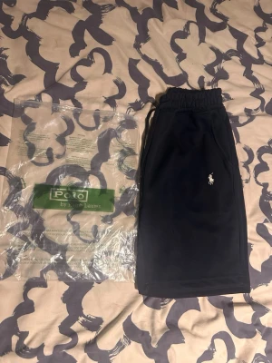 Svarta shorts från Polo Ralph Lauren - Snygga svarta shorts från Polo Ralph Lauren med klassisk logga broderad på vänster ben. Shortsen har elastisk midja med snörning och sidofickor. Perfekta för en avslappnad och stilren look.