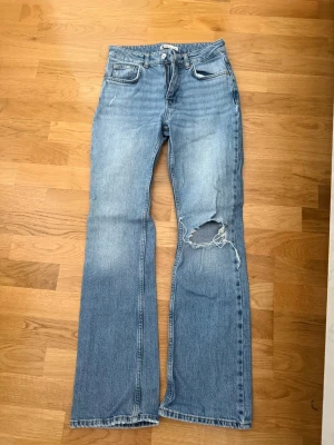 Blå bootcut jeans med slitning - Säljer ett par ljusblå bootcut jeans med slitning och hål på höger knä. Jeansen har klassisk femficksdesign, normal passform och är tillverkade i bomull. Perfekta för en avslappnad och trendig look.