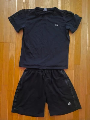 Svart set (t-shirt+shorts) från Calvin Klein - Svart set (t-shirt+shorts) från Calvin Klein med liten vit ck-logga på bröstet. Klassisk rund halsringning och korta ärmar. Logga även längs kanter på shorts och ärmar. Tillverkad i mjuk bomull som känns skön mot huden. Perfekt för en clean och enkel stil.