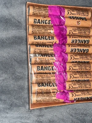Banger smällare med lila topp - Ett paket med bruna Banger-smällare med lila toppar. Smällarna är cylindriska och förpackade i plast. Märkningen och texten är tryckt i svart på sidan av varje smällare.