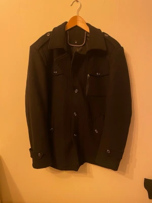 Svart rock med knappar och fickor - Stilren svart rock med klassisk krage, knäppning framtill och flera fickor, inklusive en bröstficka med dragkedja. Rocken har axelklaffar och dekorativa knappar vid ärmsluten. Perfekt för dig som vill ha en tidlös och snygg look.   (Liten XL)