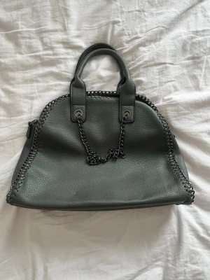 Mörkgrå handväska med kedjedetaljer från italiensk marknad  - Cool mörkgrå handväska i läderimitation med kedjedetaljer längs kanterna och en kedja som axelband. Väskan har två stadiga handtag och en dragkedja på baksidan. Perfekt för dig som gillar en edgy och stilren look. 