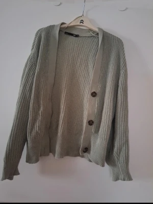 Stickad grön kofta från 157 - Säljer en stickad kofta i ljusgrön nyans från 157. Koftan har tre stora bruna knappar framtill, ribbade muddar och ett klassiskt mönster. Perfekt för lager-på-lager och att slänga över en t-shirt. Enkel och stilren design som passar till många outfits. Såg att tråden är lite uppdragen på ett ställe men inget som stör. 