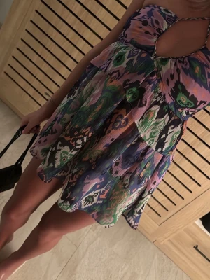 Färgglad kort klänning med mönster - Kort klänning med tunna axelband och färgstarkt, abstrakt mönster i blått, grönt, rosa, lila och vitt. Klänningen har en v-ringad urringning med twistdetalj framtill och luftig passform. Perfekt för varma dagar och sommarens alla äventyr.