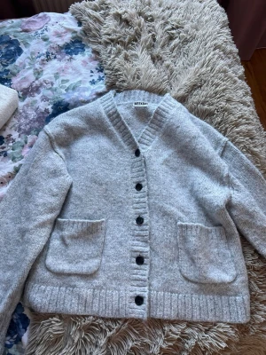 Ljusgrå kofta från Weekday - Mysig ljusgrå kofta från Weekday med v-ringad hals, stora framfickor och svarta knappar. Koftan är stickad i ett mjukt material och har ribbade muddar vid ärmslut och nederkant. 