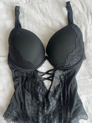 Svart spetsbody - Snygg svart body med push-up kupor och spetsdetaljer. Axelbanden är justerbara och framsidan har ett dekorativt snörningsmönster. Materialet är en kombination av mjuk mesh och spets som ger en elegant look.