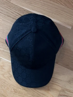 Svart Gucci keps med GG-mönster - Snygg svart keps från Gucci med det klassiska GG-monogrammet över hela kepsen. På sidorna finns gröna och röda ränder som ger en ikonisk touch. Kepsen har böjd skärm och justerbar passform. Materialet är troligtvis bomull eller en bomullsblandning. Helt ny