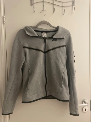 Grå zip hoodie från Nike - Snygg grå hoodie från Nike med dragkedja framtill och svarta detaljer. Tröjan har huva, två fickor med dragkedja och en liten ficka på ärmen. Tillverkad i mjukt sweatshirtmaterial, perfekt för en sportig och avslappnad stil.