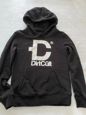 Svart hoodie från DirtCult - Snygg svart hoodie från DirtCult med stor vit logga och text på bröstet. Klassisk känguruficka framtill, huva med dragsko och ribbade muddar. Perfekt för dig som gillar streetwear och vill ha en enkel men stilren look.