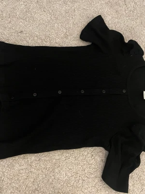 Svart ribbad topp från Lindex - Säljer en svart ribbad topp från Lindex med korta puffärmar och knappar framtill. Toppen har en rund halsringning och är tillverkad i mjuk bomull, perfekt för en enkel men snygg look.