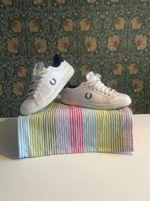 Vita Fred Perry sneakers med svart detalj -  Vita sneakers från Fred Perry med klassisk lagerkrans-logga på sidan och bak. Skorna har lågt skaft, och vita snören. Perfekta för en clean och stilren look. Liten skada fram på ena skon 