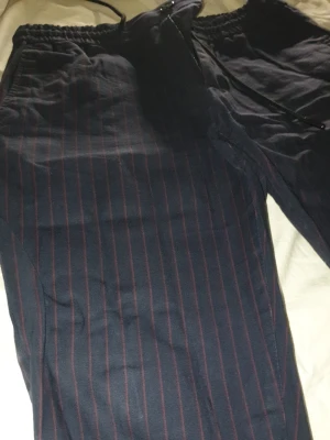 Mörkblå randiga mjukis/kostym byxor - Säljer ett par mörkblå mjukis/kostym byxor med tunna röda ränder. Byxorna har resår i midjan och dragsko samt fickor på sidorna och en bakficka. Sköna och avslappnade, perfekta för chill dagar. Materialet känns mjukt och bekvämt.