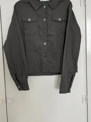 Svart jeansjacka med silvriga knappar - Säljer en stilren grå jacka/skjortjacka i fint skick. Den är bekväm att bära och passar perfekt både till vardag och mer uppklädda outfits. En tidlös modell som är lätt att matcha med olika plagg.  Jackan har krage, knäppning framtill med knappar samt två bröstfickor som ger en snygg och klassisk look. Mjukt och skönt material som sitter fint på kroppen.  Hör gärna av dig vid frågor eller om du vill se fler bilder!