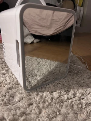 Vitt sminkkylskåp med spegeldörr - Snyggt och modernt sminkkylskåp i vitt med spegeldörr och praktiskt handtag upptill. Kylskåpet har en hylla och en låda för smart förvaring av hudvård och smink. Tillverkat i plast med spegelyta på dörren, perfekt för ditt sminkbord.