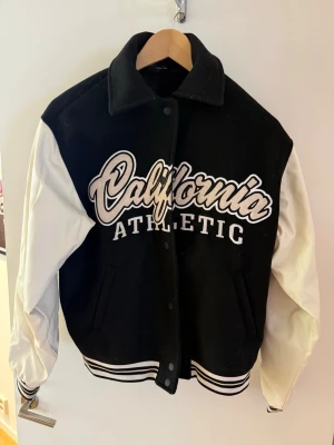Svart och vit varsityjacka California - Snygg svart och vit varsityjacka med stora broderade bokstäver 'California Athletic' på bröstet. Jackan har vita ärmar, svarta muddar med vita ränder och klassisk collegekrage. Perfekt för en sportig och trendig look.