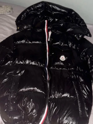 Moncler montbeliard, Äkta, pris kan diskuteras  - Säljer en svart, glansig pufferjacka från Moncler med huva och klassisk logga på bröstet. Jackan har dragkedja med röd, vit och blå detalj längs öppningen och stora fickor framtill. Perfekt för kalla dagar och riktigt snygg streetstil.