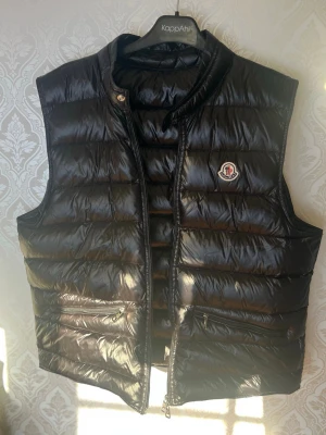 Moncler väst - Snygg svart dunväst från Moncler med glansig finish och klassisk quiltad design. Äktenhetskap kan såklart visas genom kod och scanner på både nfc scanner och innuti✅🧾.