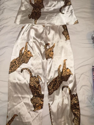  pyjamas med leopardmönster - Snygg tvådelad pyjamas i silvrigt satinmaterial med coolt leopardmönster. Setet består av ett linne med smala axelband och ett par lösa byxor, båda med stora leopardtryck i brunt och svart. Perfekt för dig som vill ha något extra till kvällsmyset.