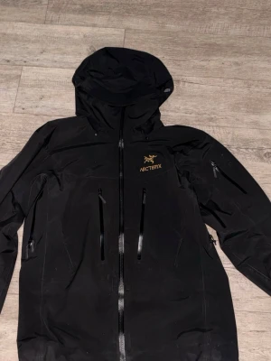 Svart vindjacka från Arc'teryx - Snygg svart vindjacka från Arc'teryx med huva och dragkedjor framtill. Jackan har två vertikala bröstfickor med vattentäta dragkedjor och broderad logga i gult på bröstet. Perfekt för blåsiga dagar och outdoor-äventyr.