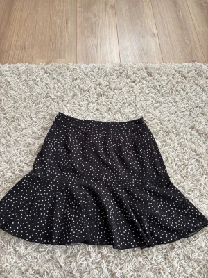 Svart prickig kortkjol med dragkedja - Snygg svart kortkjol med vita prickar och en diskret dragkedja bak. Kjolen har en lätt utsvängd form och är perfekt för dig som gillar en lekfull men stilren look. Tillverkad i ett mjukt och följsamt material.