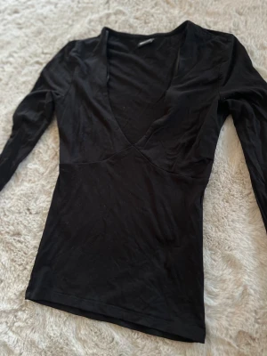 Svart långärmad v-ringad topp - En svart långärmad topp med djup v-ringning och figurnära passform. Perfekt för att skapa en snygg och enkel look. Materialet känns mjukt och stretchigt, vilket gör den bekväm att bära hela dagen.