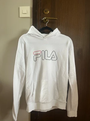 Vit hoodie från FILA med logga - Snygg vit hoodie från FILA med stor logga i svart och rött framtill. Klassisk känguruficka och huva. Tillverkad i mjukt bomullsmaterial som är skönt att ha på sig. Säljer för att den är för liten. Storlek 158-164. 
