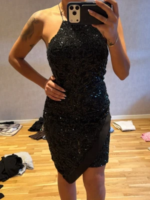 Svart glittrig kort klänning - Säljer en svart glittrig kort klänning med halterneck och figurnära passform. Klänningen har ett snyggt mönster med paljetter och en diagonal detalj framtill. Perfekt för dig som vill sticka ut på festen!